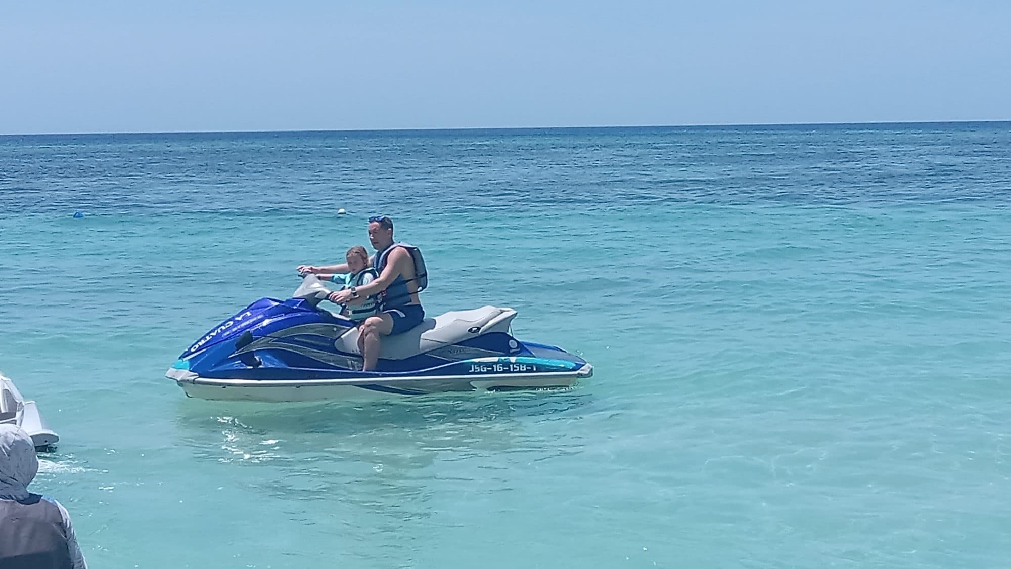 Un adulto conduciendo una moto de agua azul con un niño pequeño sentado delante de él en agua clara y poco profunda