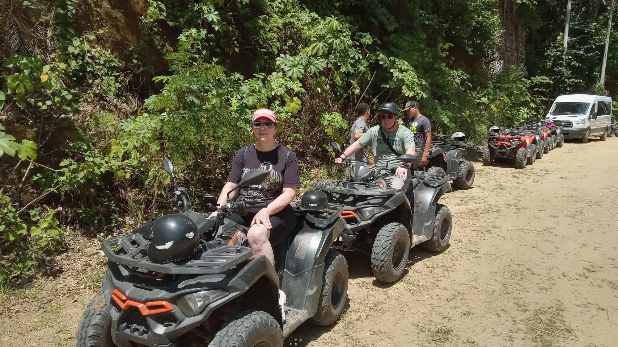 Dos personas en cuatrimotos al frente de una fila de quads en un camino de tierra entre la selva, con la van del tour aparcada detrás