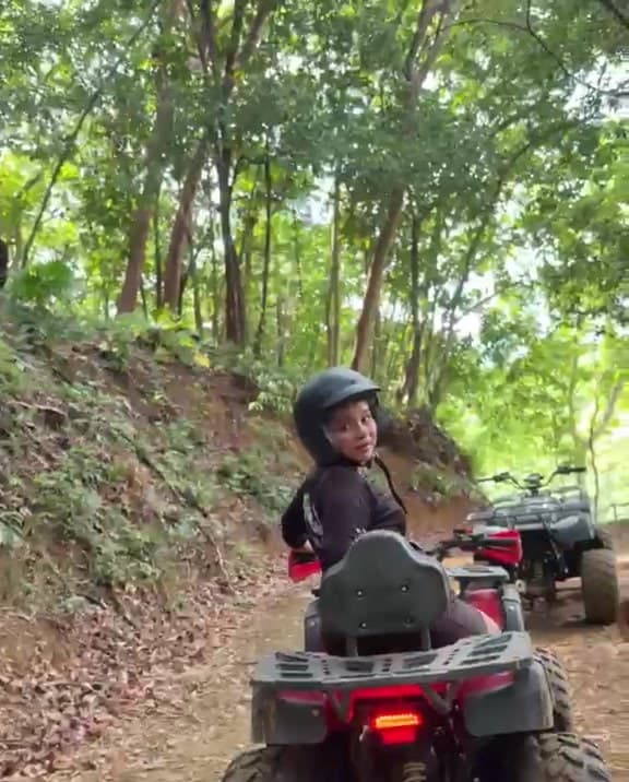 Una visitante con casco subiendo por un camino de tierra en cuatrimoto entre la selva, con un guía caminando al lado