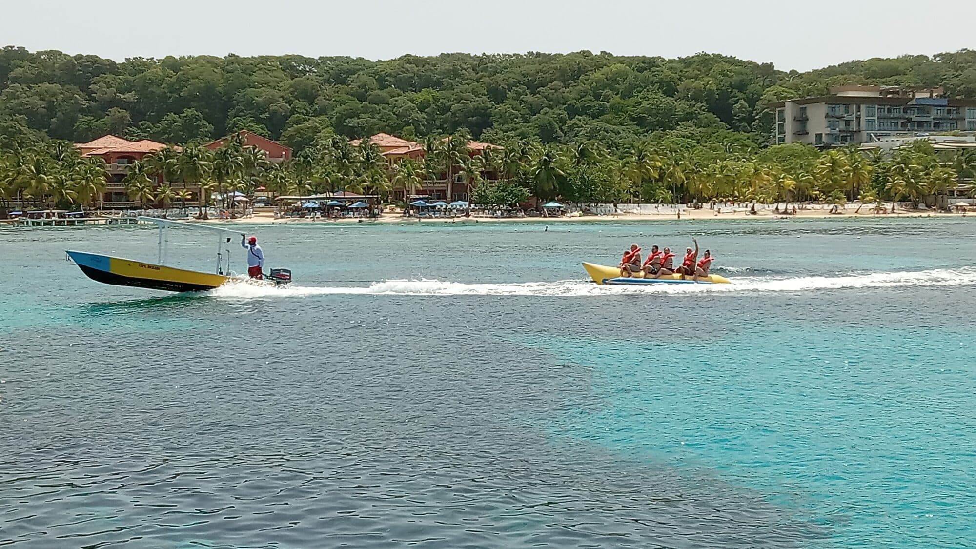 Seis personas con chalecos rojos en un banana boat amarillo remolcado por la bahía, con la playa y las palmeras al fondo