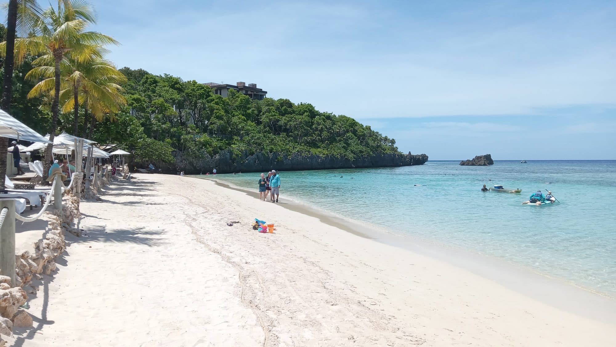 Una playa blanca y estrecha que se curva bajo un acantilado de roca caliza cubierto de selva, con una familia a la orilla