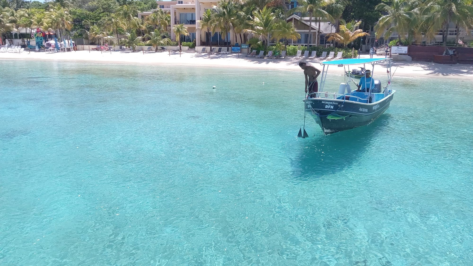 Una lancha de tour fondeada en agua tan clara que se ve el fondo de arena, con un tripulante en la proa y el resort detrás