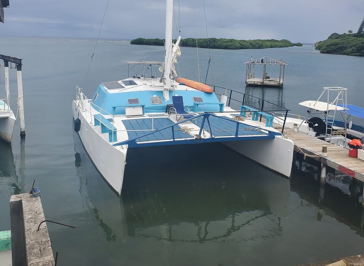 Un catamarán azul y blanco amarrado a un muelle en una bahía tranquila, con islotes de manglar en el horizonte