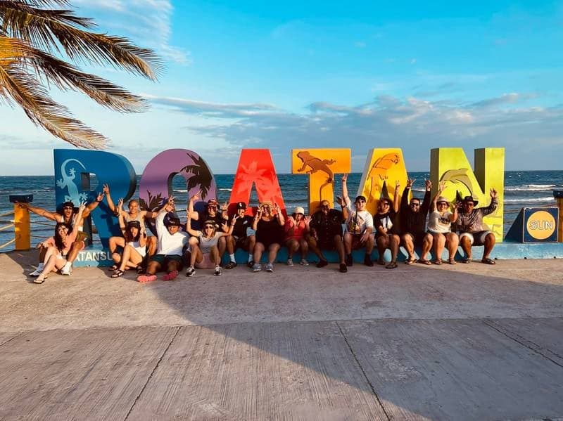 Una veintena de visitantes sentados a lo largo de las letras gigantes de ROATÁN junto al mar al atardecer, con los brazos en alto
