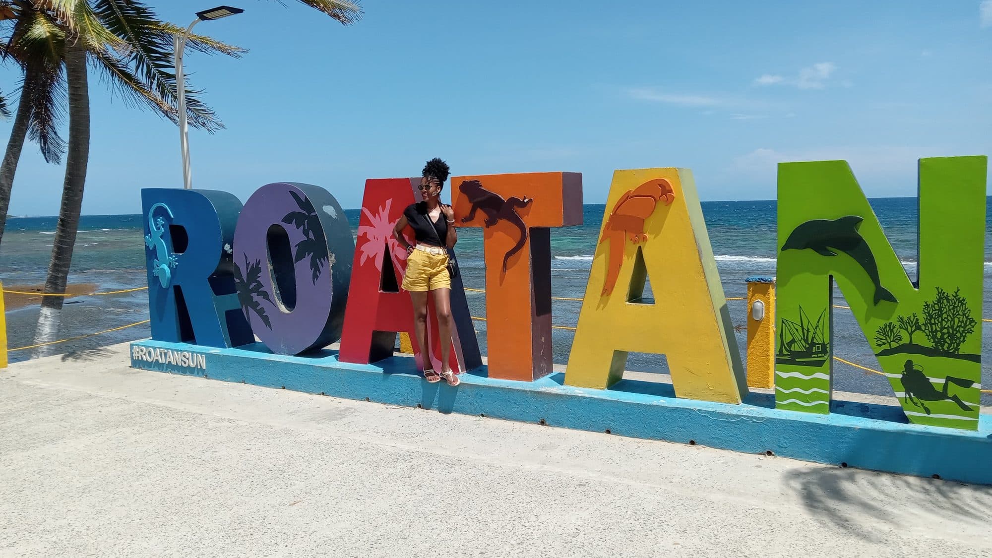 Una visitante posando entre las letras gigantes pintadas de ROATÁN con el mar detrás