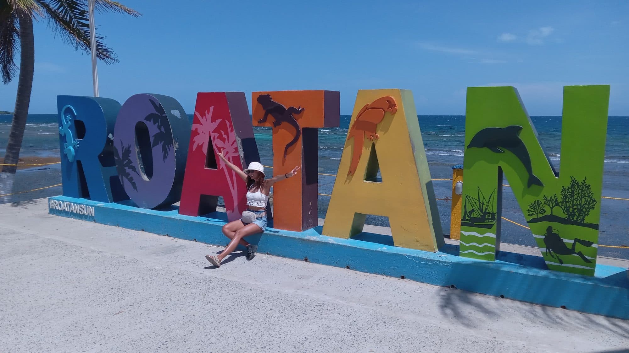 Una visitante sentada en el zócalo azul, en el hueco entre dos de las letras gigantes pintadas de ROATÁN, con el mar detrás