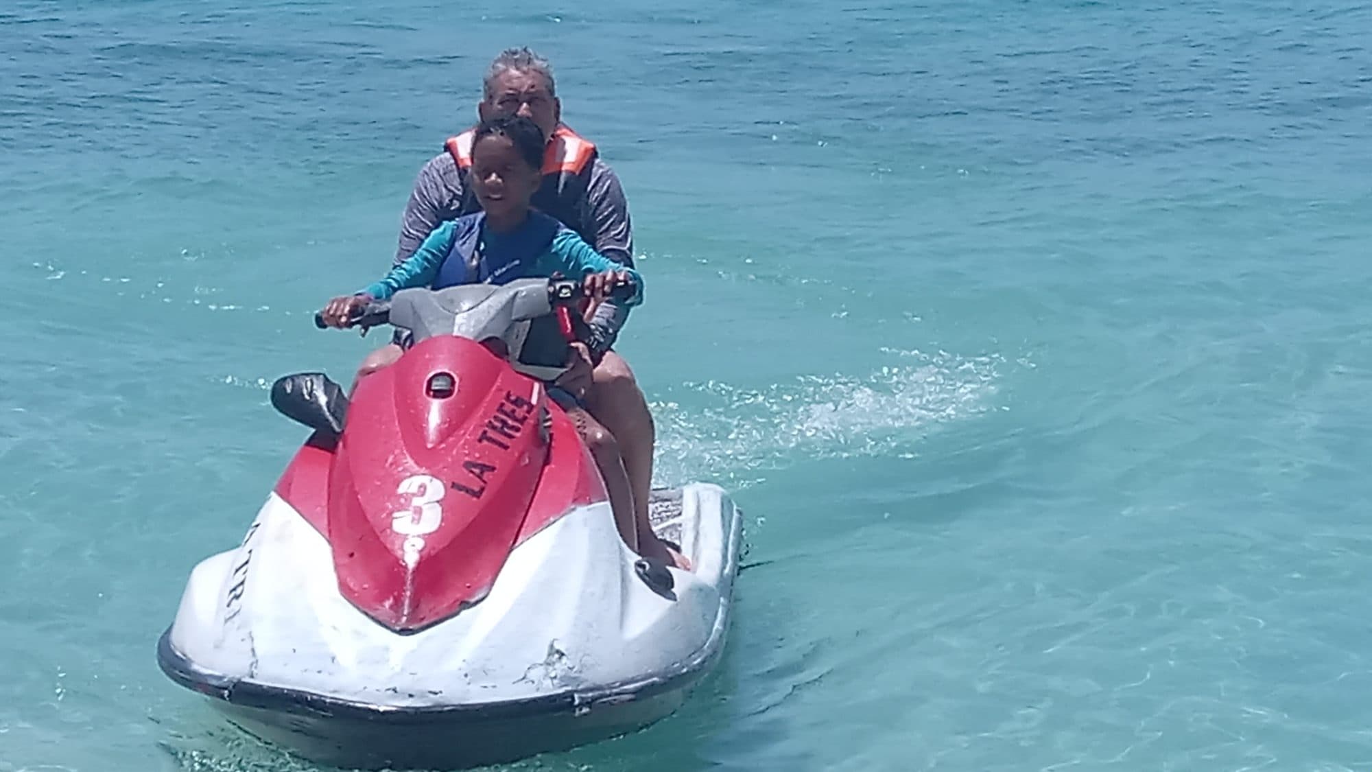 Dos visitantes con chaleco salvavidas sentados en una moto de agua roja y blanca a la orilla, listos para salir
