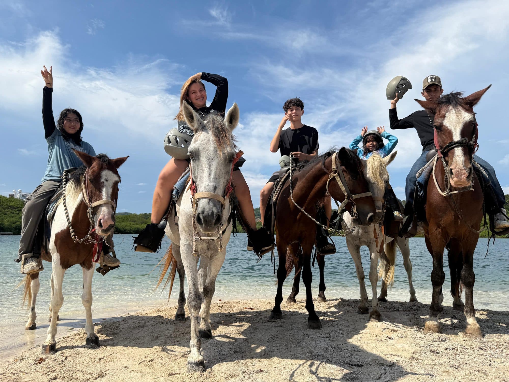 Cinco jinetes a caballo al borde de la playa, varios con los brazos en alto y el mar detrás