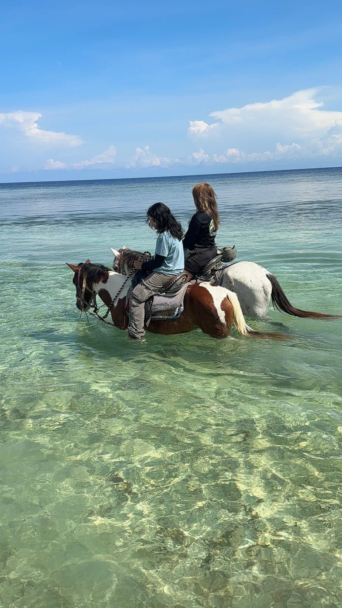 Two riders side by side on horses standing in clear shallow water under a blue sky