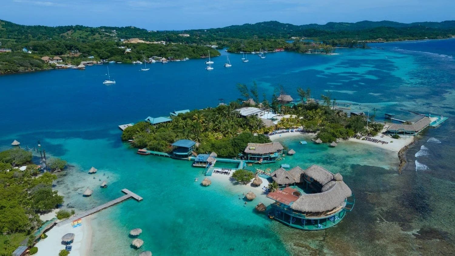 Little French Key desde el aire: la isla privada con sus cabañas de palma sobre el agua, la plataforma clara del arrecife alrededor y veleros fondeados en la bahía del fondo