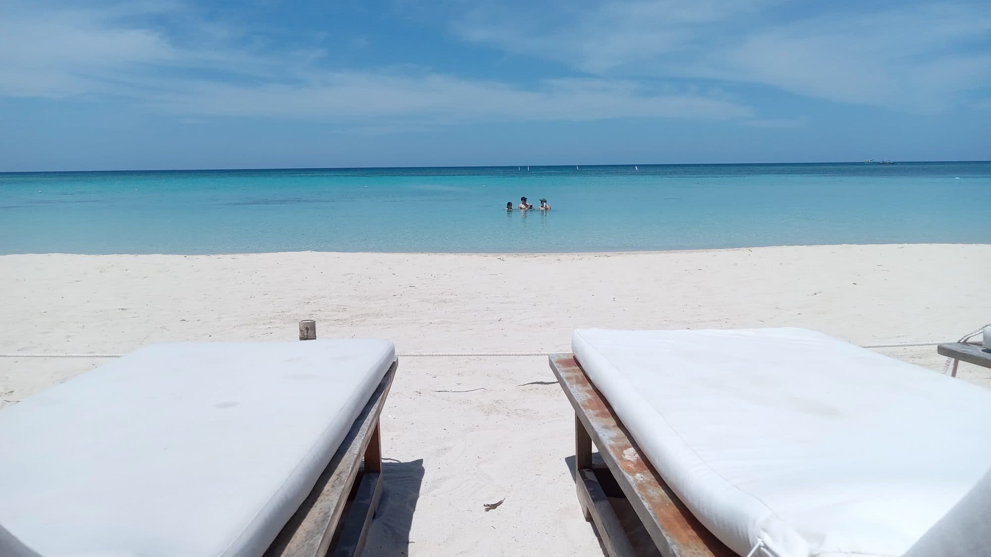 La vista por encima de dos camastros vacíos hacia el mar, con bañistas a lo lejos