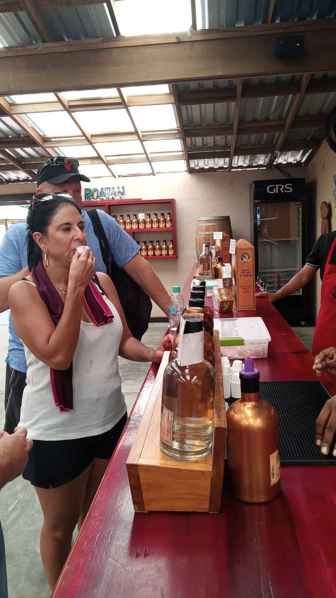 Una visitante probando ron en el mostrador de la tienda, con botellas alineadas en los estantes y un rótulo de ROATÁN en la pared