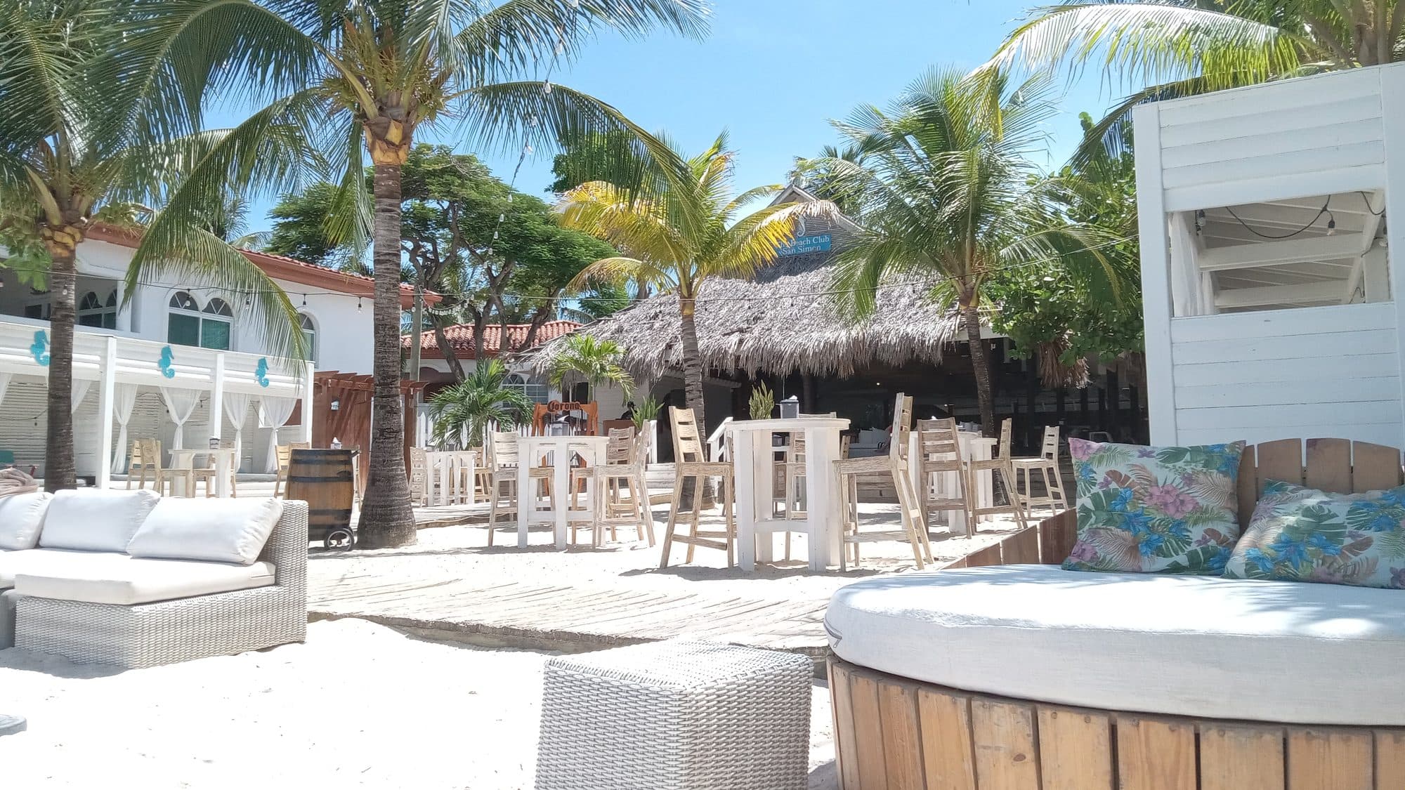 El bar de playa al aire libre bajo su techo de palma, con taburetes en la arena y una tina de madera en primer plano