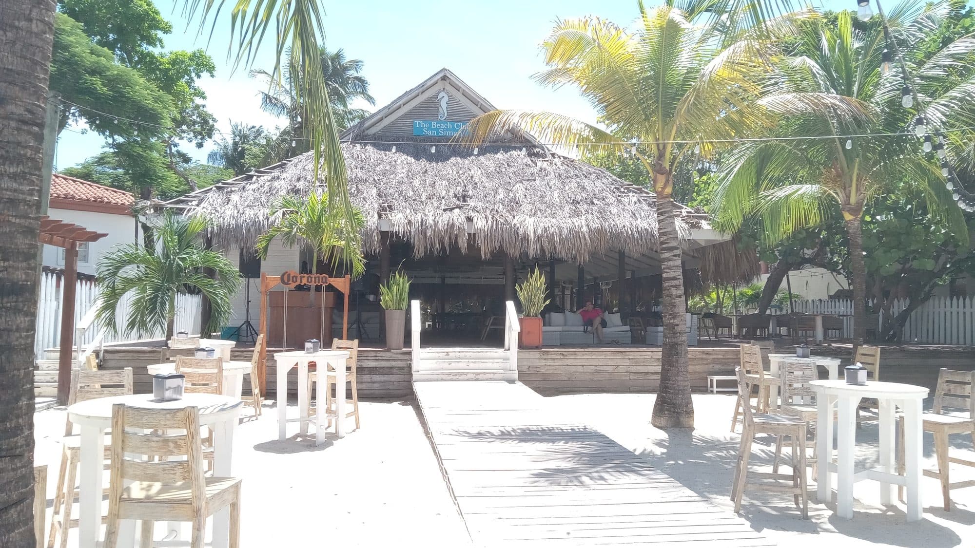 El techo de palma de The Beach Club San Simón visto desde la pasarela de madera, con su rótulo sobre la barra