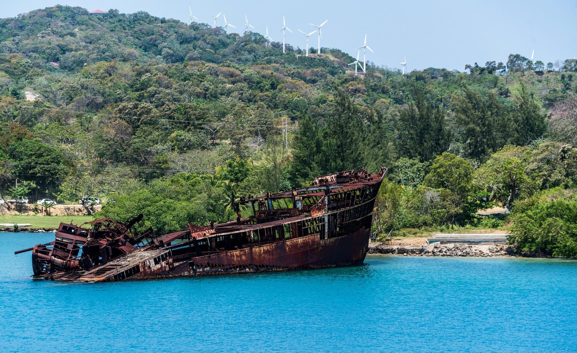 Un barco naufragado y oxidado varado contra la orilla bajo una ladera verde coronada por aerogeneradores