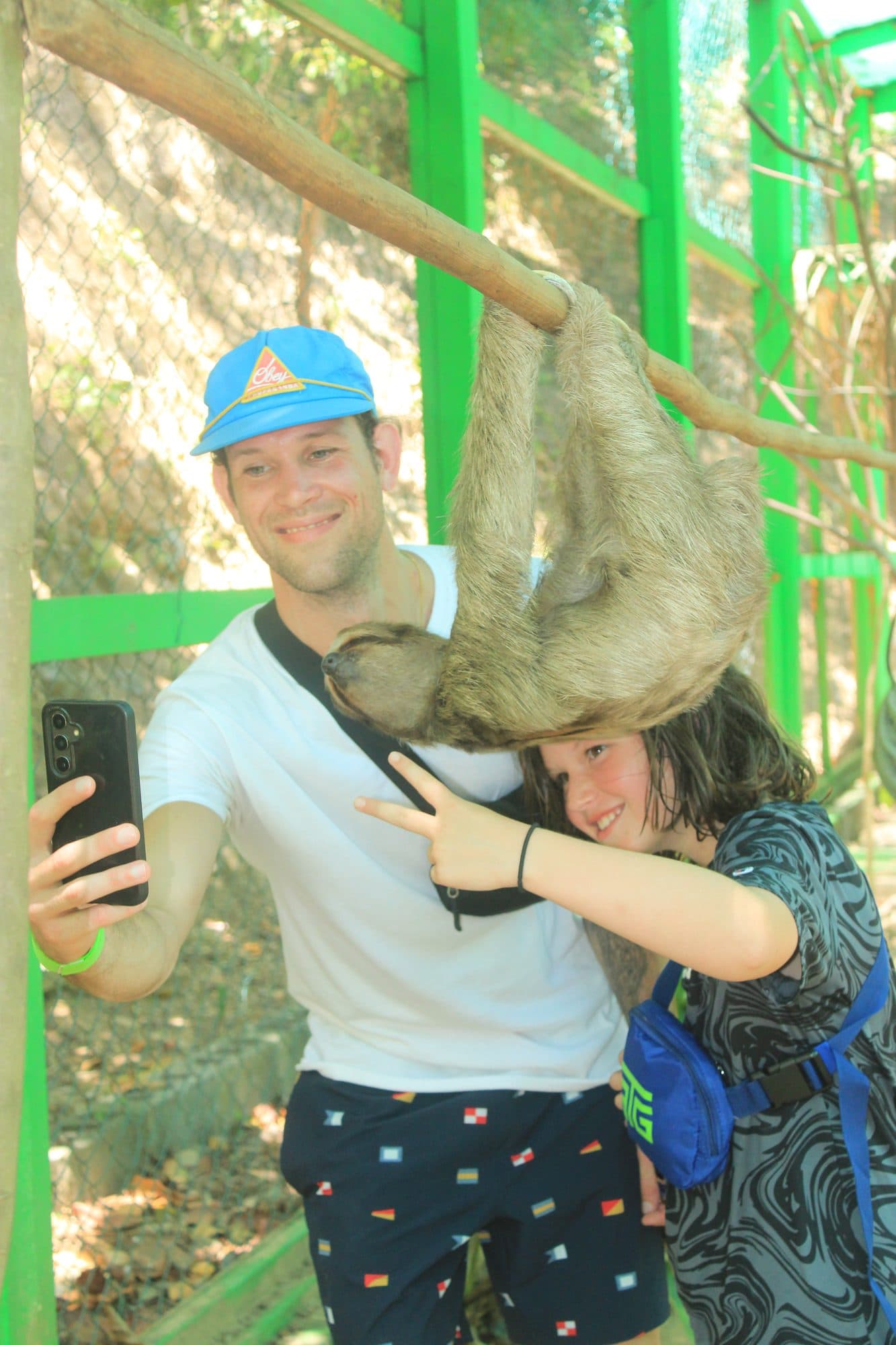 Un visitante y un niño haciéndose un selfie mientras un perezoso cuelga de la rama justo encima