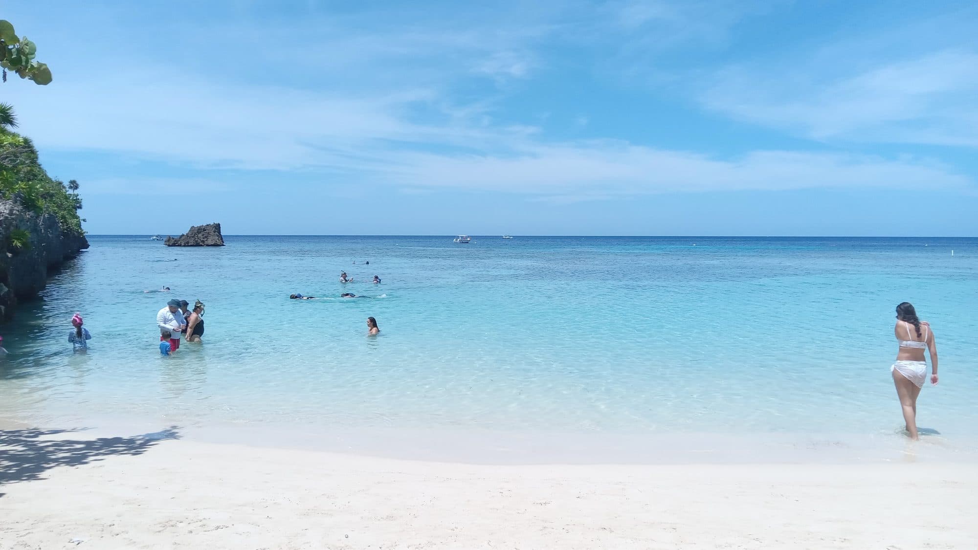 Una docena de visitantes repartidos en las aguas bajas turquesa frente a una playa blanca, unos de pie y otros boca abajo haciendo snorkel más adentro