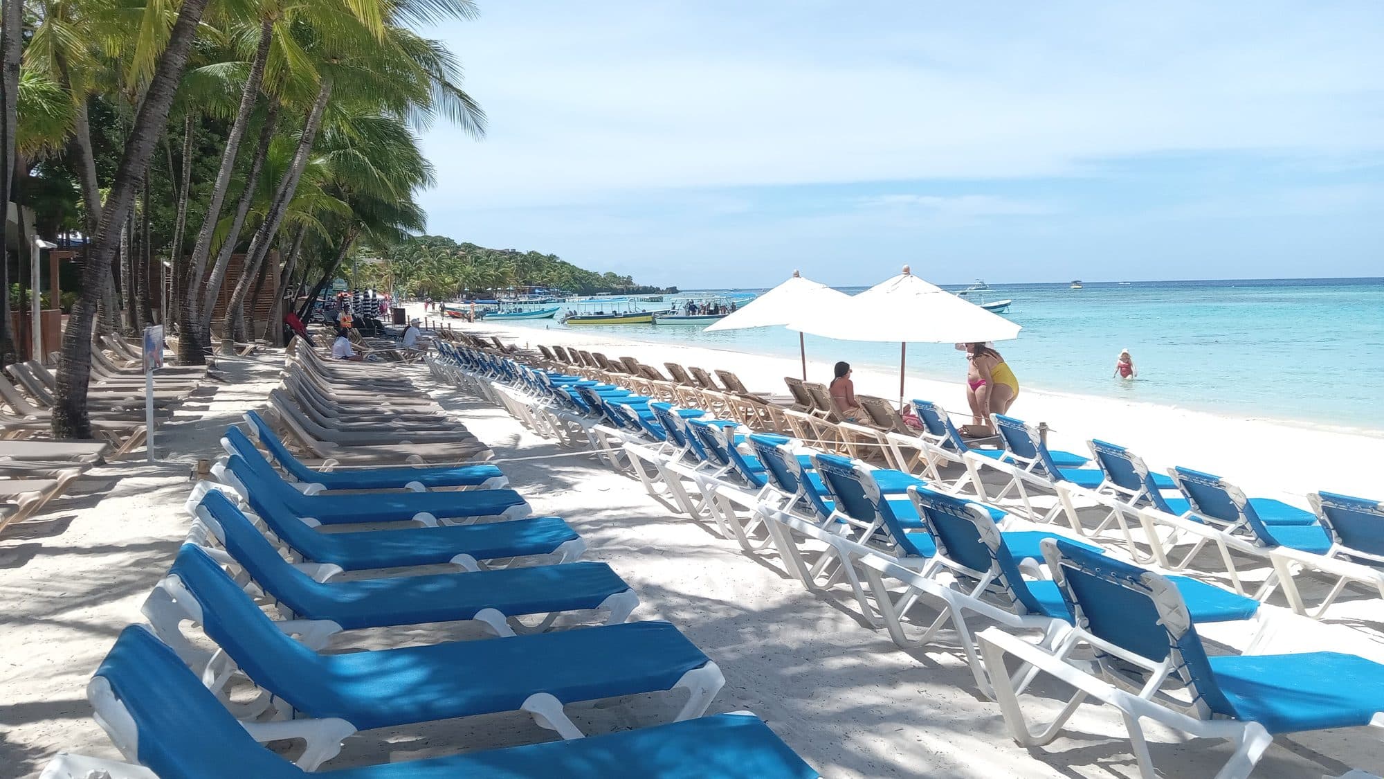 Una fila larga de camastros azules y sombrillas blancas en Playa West Bay con visitantes sentados al sol