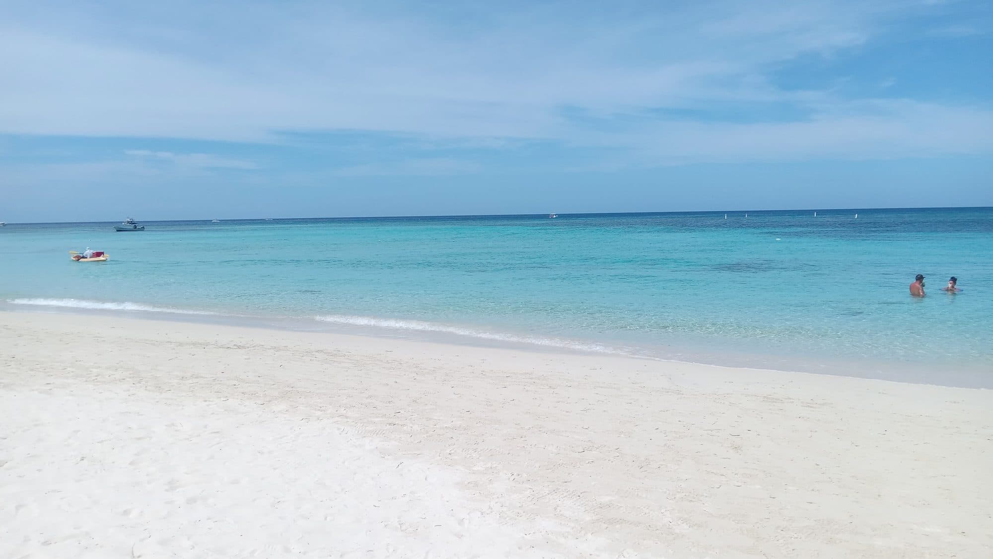 Arena blanca ancha en West Bay con dos bañistas y una lancha pequeña en el agua turquesa en calma