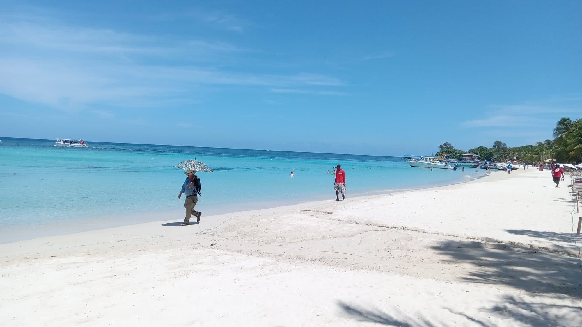 Dos personas caminando por la orilla vacía de West Bay, una con una sombrilla, con el agua en calma hasta el horizonte